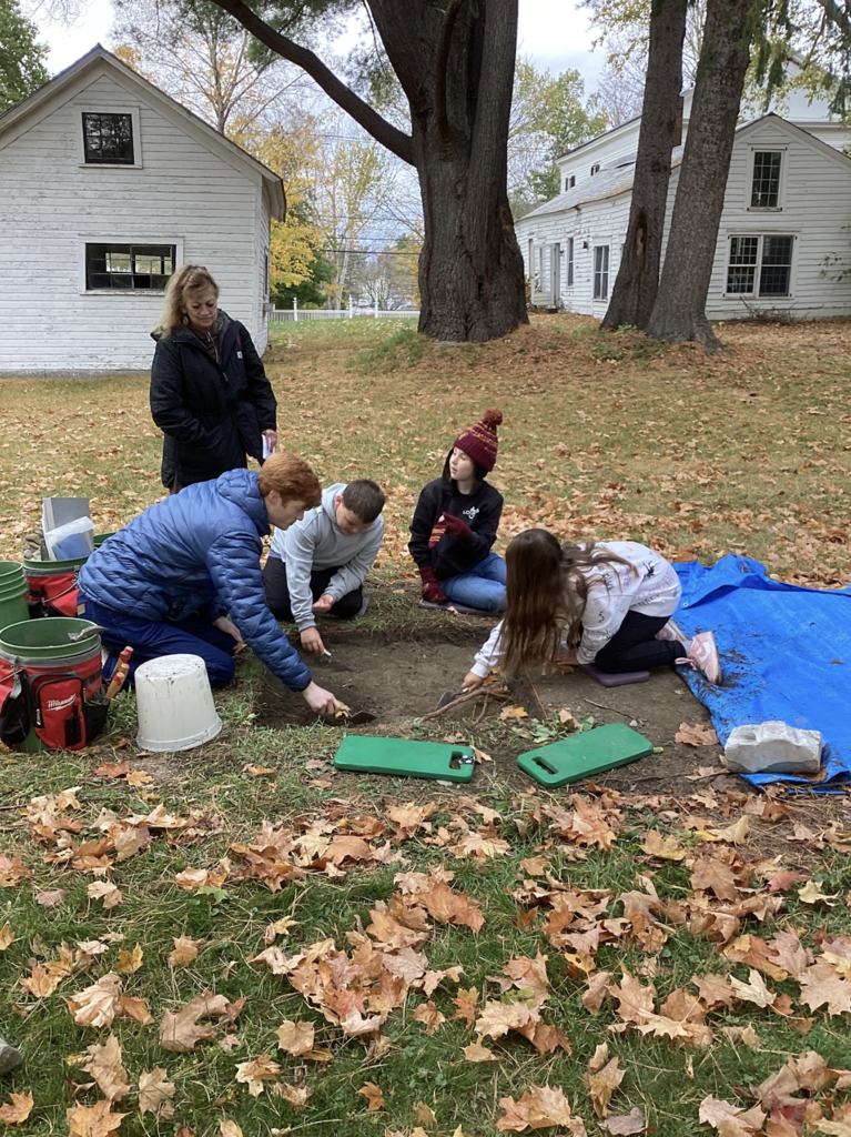Students and a teacher dig and explore an excavation site surrounded by autumn leaves and old buildings.