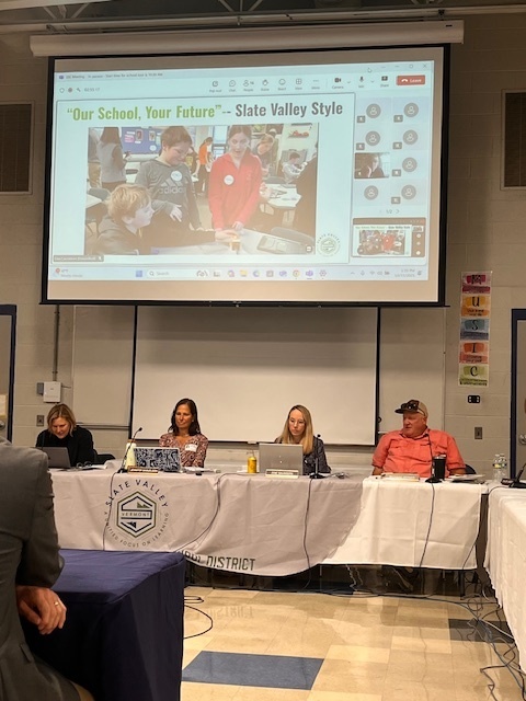 A presentation titled “Our School, Your Future – Slate Valley Style” is projected behind panelists at a district meeting.