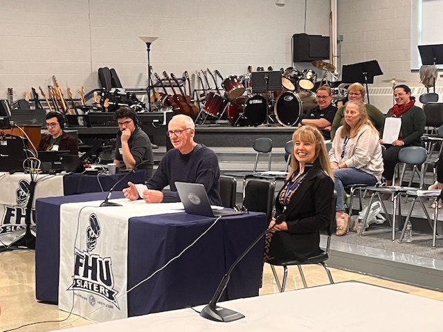 Two presenters at a school board meeting share updates while smiling, with instruments in the background.