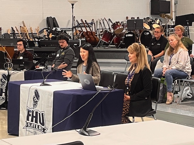 Presenters speak at a meeting table with FHU Slaters banners while attendees and equipment are visible behind them.
