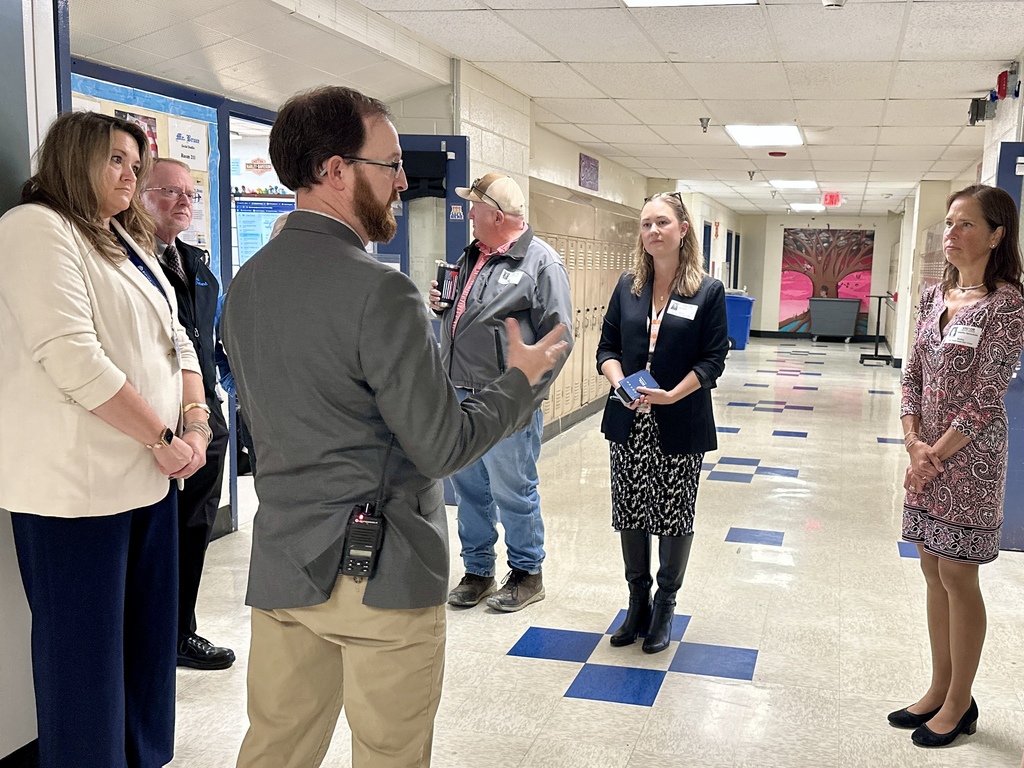 A principal leads a group of visitors through a school hallway, discussing programs and facilities.