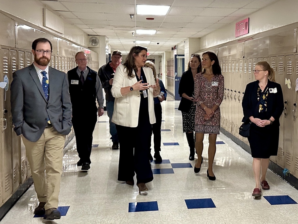Administrators and visitors walk down a hallway lined with lockers during a school tour.