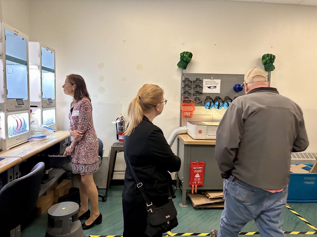 Visitors observe equipment in a classroom featuring 3D printers and safety gear on display.