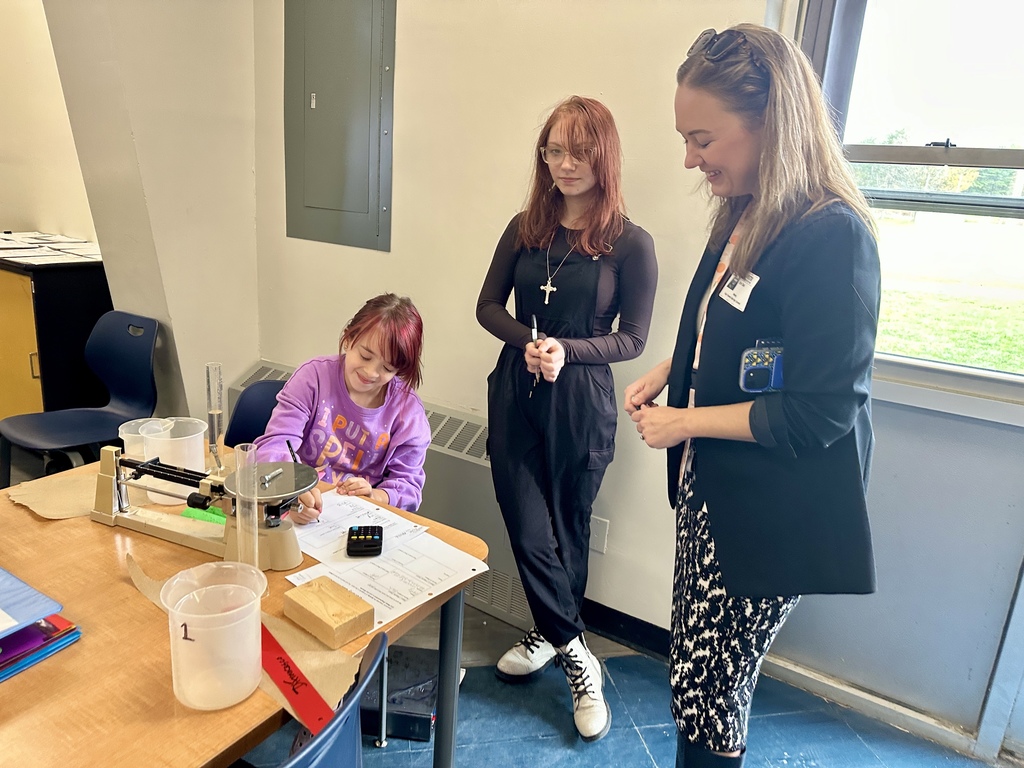 Two students work on a science experiment while a visitor smiles and engages with them.