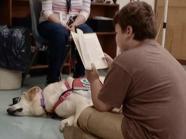 A student sits on the floor reading aloud from a book to a therapy dog resting nearby with its handler seated behind them.