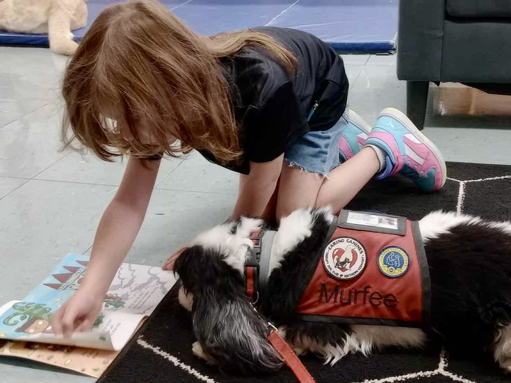 A young student kneels on the floor reading a book to a therapy dog named Murfee, who is lying beside her wearing a red service vest.