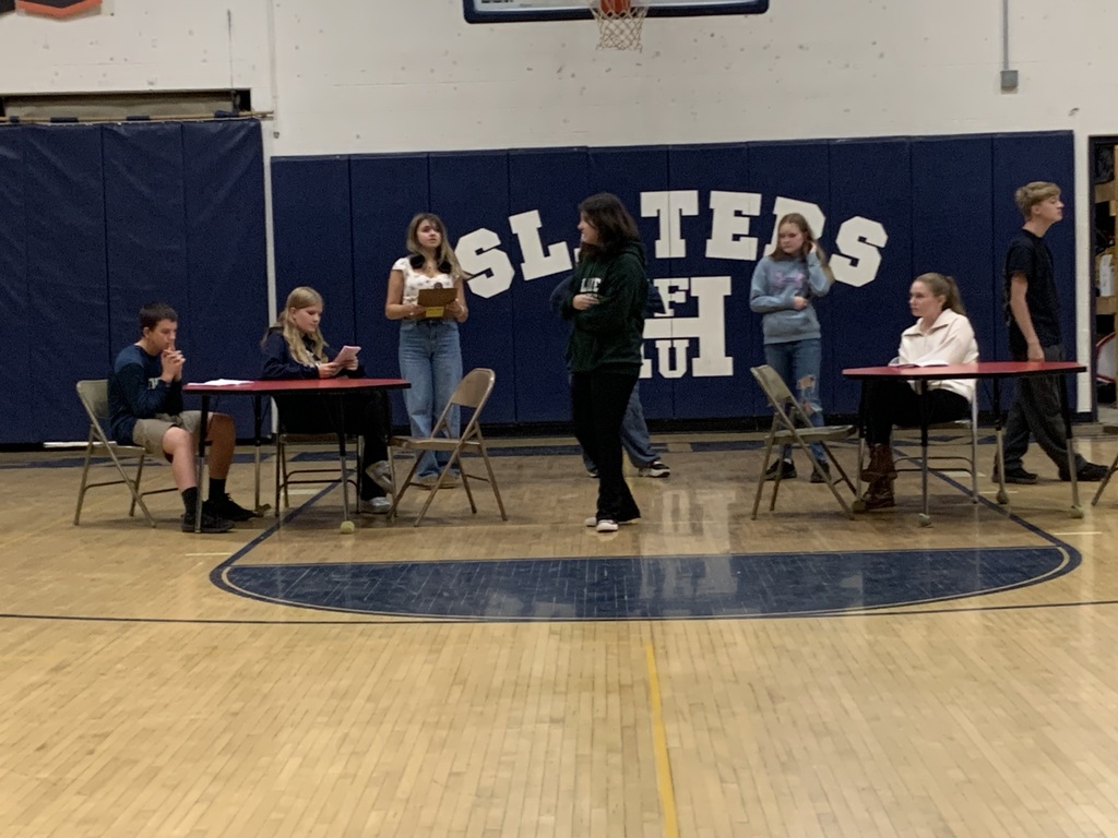 Students perform a classroom skit in the gymnasium, standing and sitting at tables in front of a “Slaters” wall backdrop.
