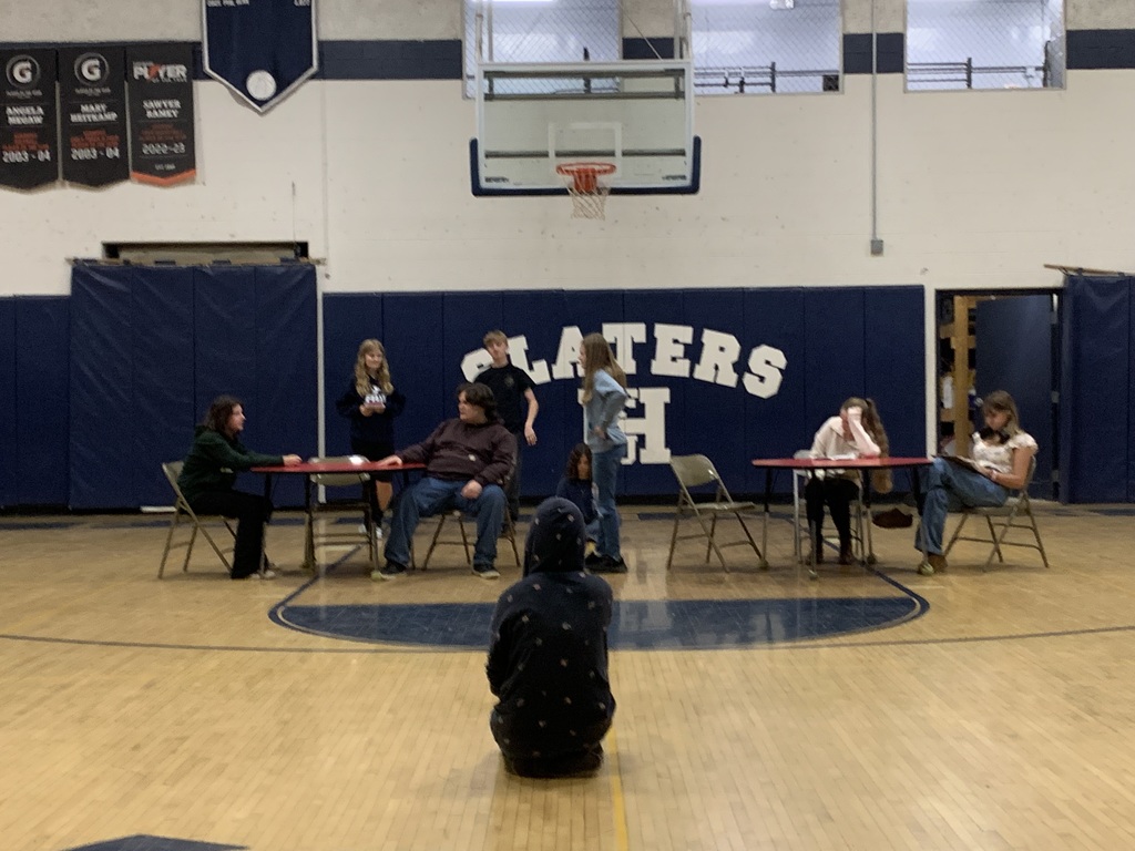 A group of students act out a scene at tables in the gym, with one student sitting in the foreground watching the performance.