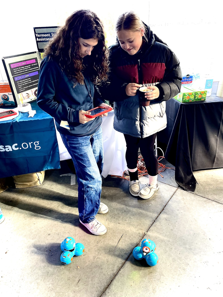 Two students control small blue coding robots using a tablet at a booth.