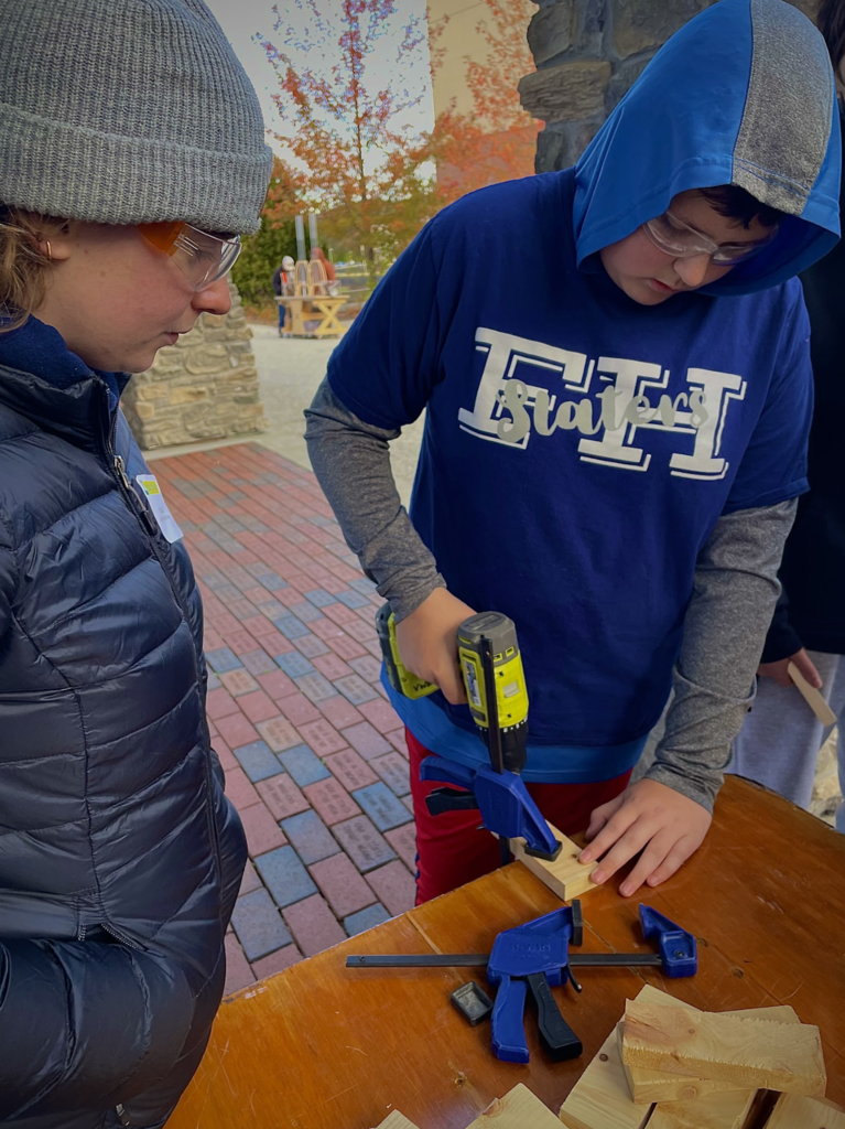 A student in safety goggles drills a piece of wood while another observes closely.