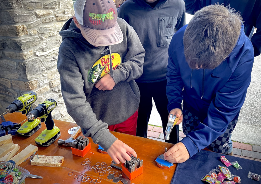 Two students work with metal stamping tools at a table with drills and key rings.