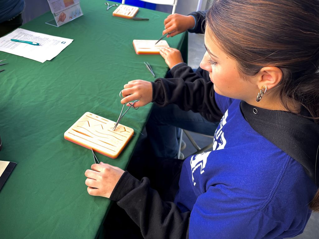 A student practices suturing on a medical training pad at a green-covered table.