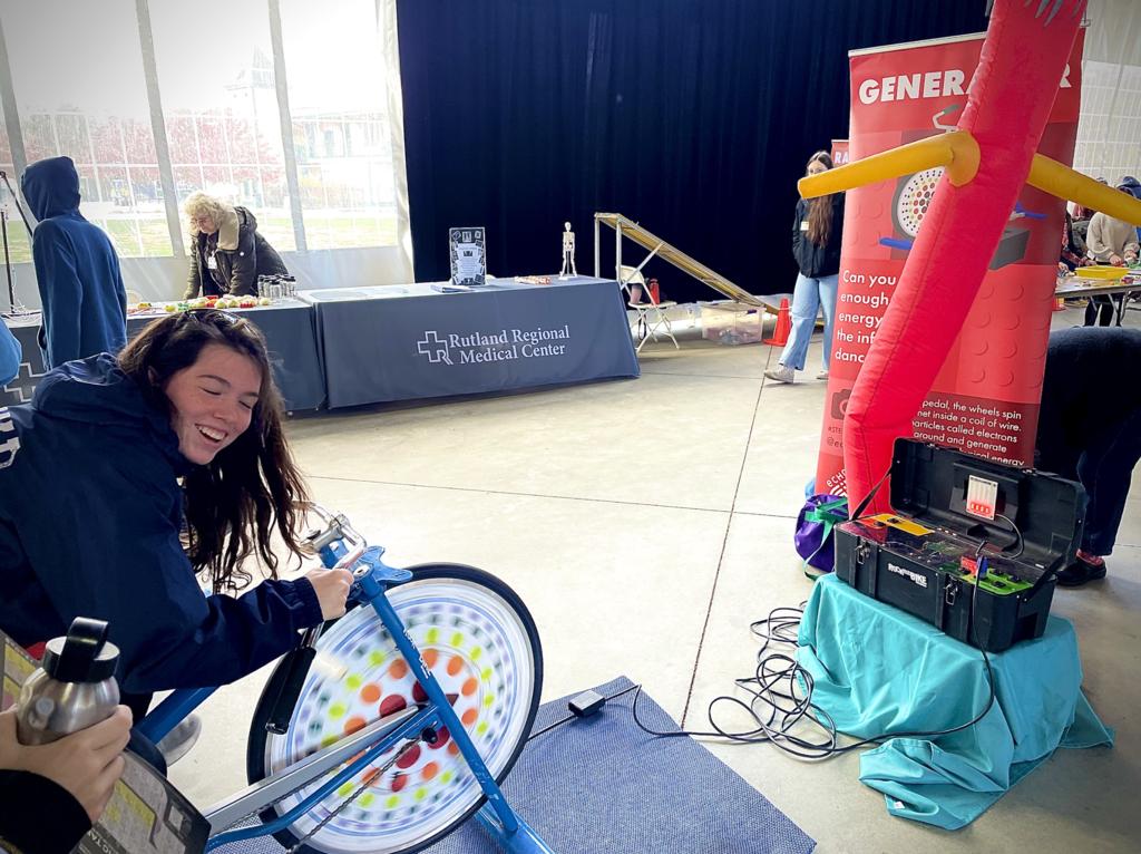A student rides a stationary bike that powers an inflatable tube display at a fair.