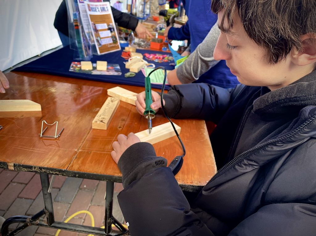 A student uses a wood-burning tool to etch on a wooden block at a workshop table.