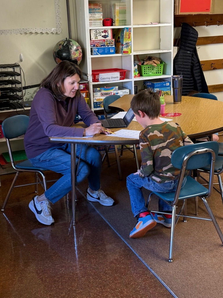 A woman (teacher or tutor) sits at a table with a young boy in a classroom, appearing to help him with schoolwork. A laptop is open on the table, and a white bookshelf filled with books and games is visible behind them
