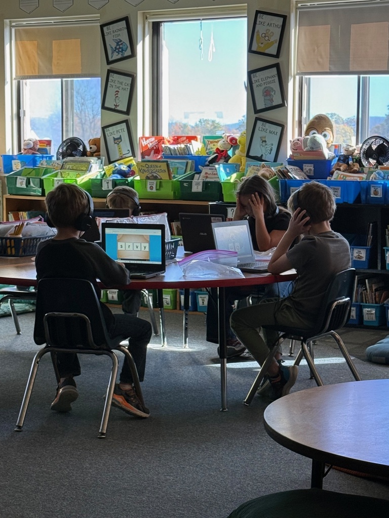 Four elementary school students sit at a red table in a classroom, each wearing headphones and working on laptops. One laptop screen shows a word-building activity. The classroom has large windows and bookshelves with books and toys.