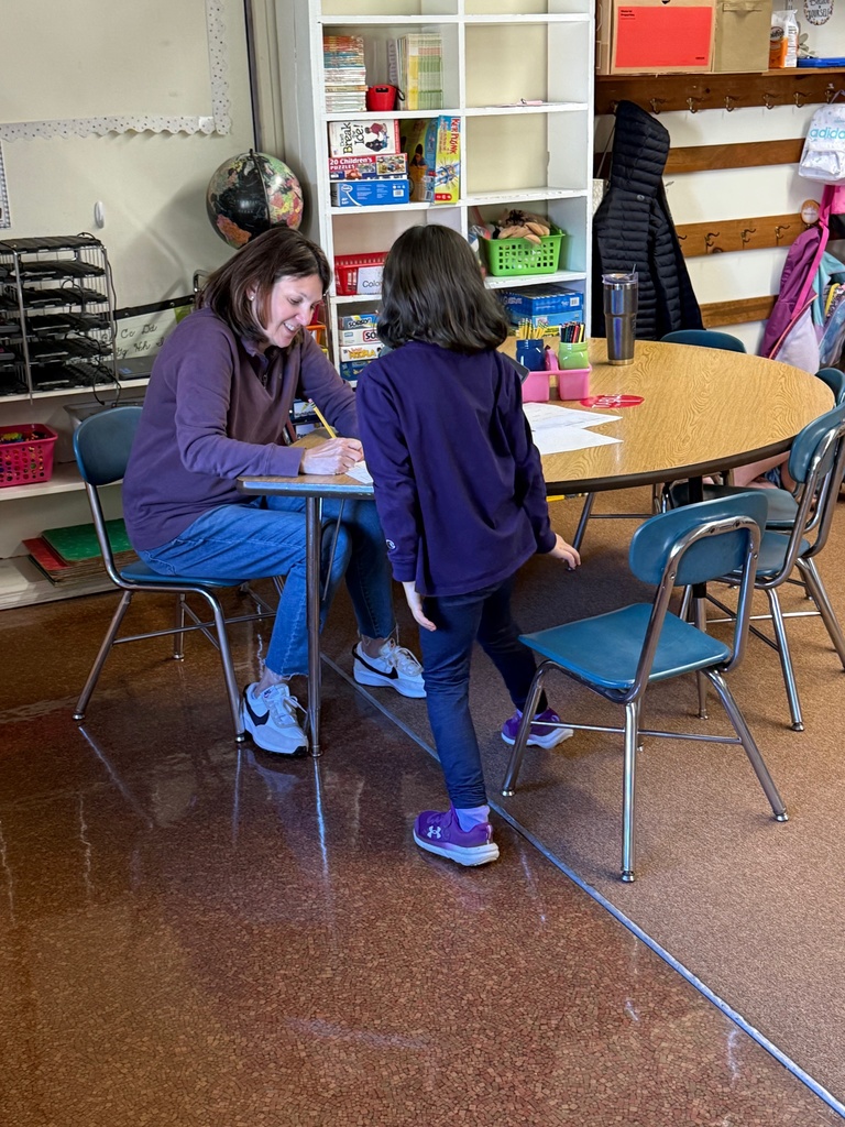 A woman (teacher or tutor) sits at a table with a young student in a classroom, appearing to help them with schoolwork. A laptop is open on the table, and a white bookshelf filled with books and games is visible behind them