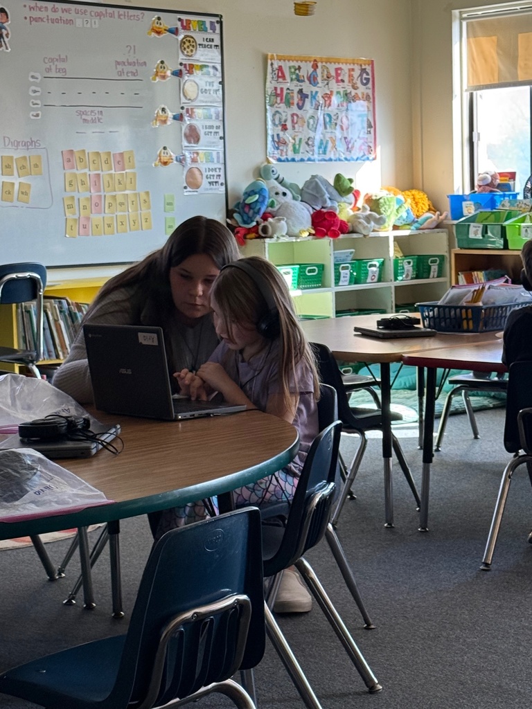 A teacher sits with an elementary school girl at a round table, helping her work on a laptop. The girl is wearing headphones. The background shows a whiteboard with phonics/literacy lessons and shelves filled with stuffed animals and classroom materials.