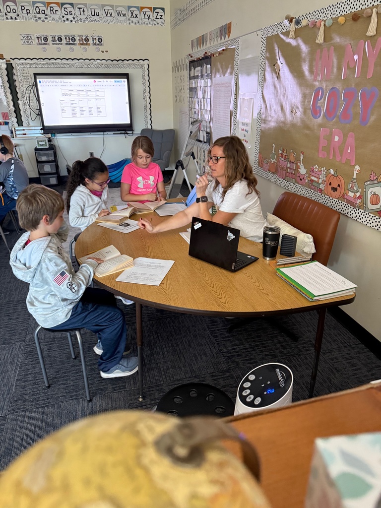 A female teacher sits at a round table with three elementary school students in a classroom, leading a small group lesson. Books, papers, and a laptop are on the table. A TV screen and an "In My Cozy Era" bulletin board are visible in the background.