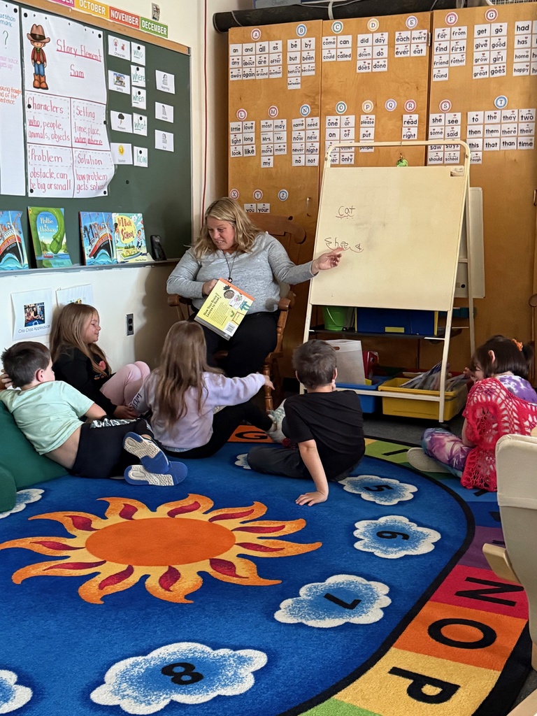 A teacher sits in a chair and reads a book to a small group of six elementary school students seated on a colorful rug in a classroom. The teacher is pointing to a word written on a small whiteboard easel. The wall is covered with educational posters and word cards.