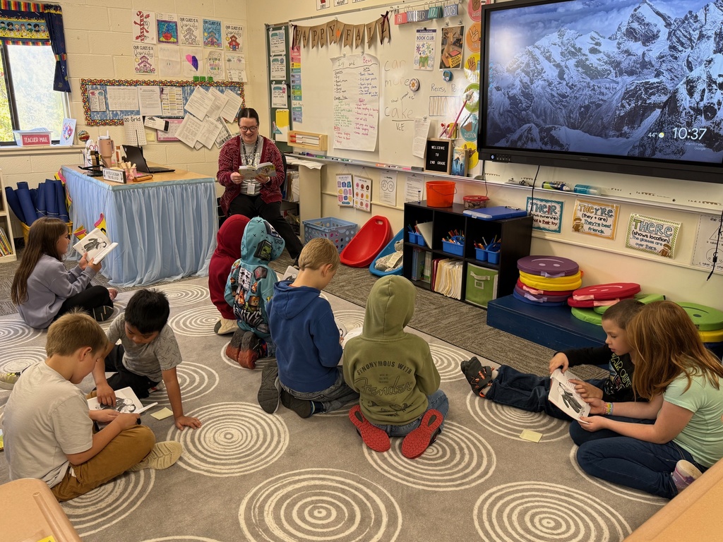 A teacher reads a book to a group of elementary school students seated on a rug in a classroom. Some students are also looking at their own books. A large screen displays a mountain range image.