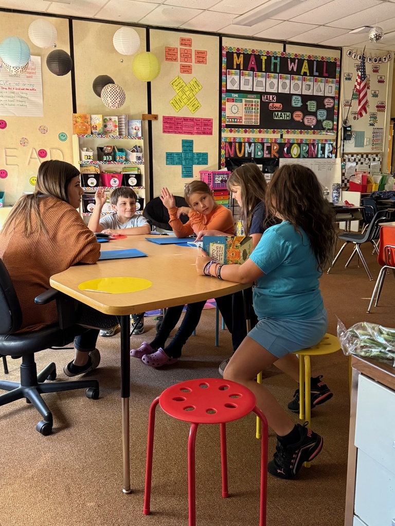 A teacher sits with four elementary school students around a small table in a colorful classroom. One boy and one girl have their hands raised, while another girl holds a book. The classroom walls are decorated with a "Math Wall" and other educational posters.
