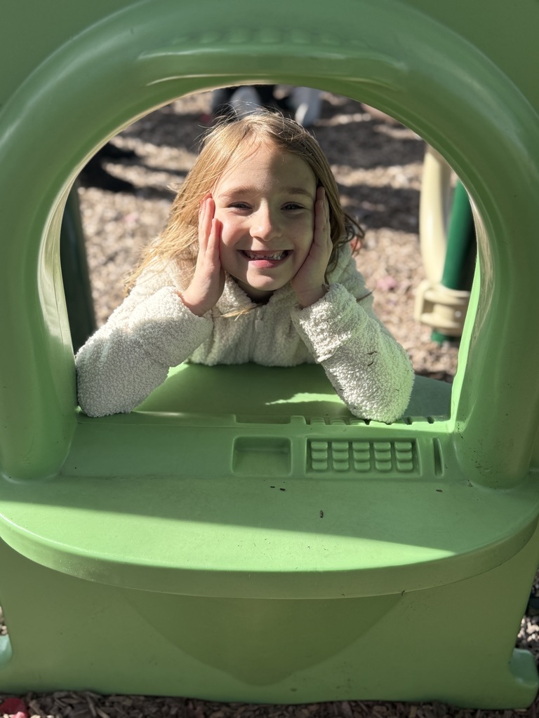 A smiling child leans through a green playground window panel.