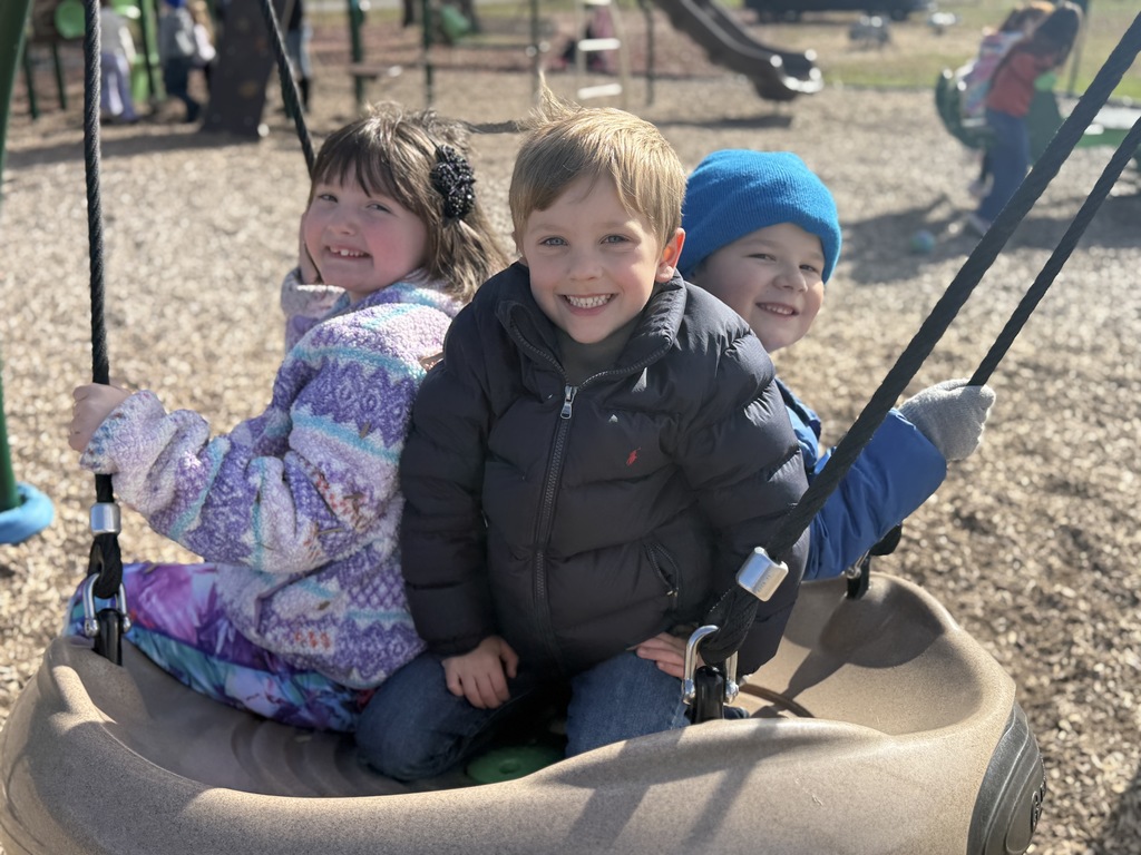 Three children sit together on a round swing, smiling on a sunny day.