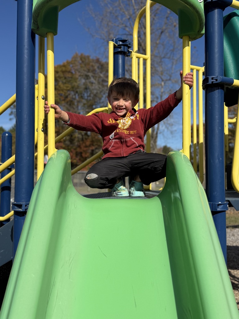 A child in a red hoodie smiles while sitting at the top of a green slide.