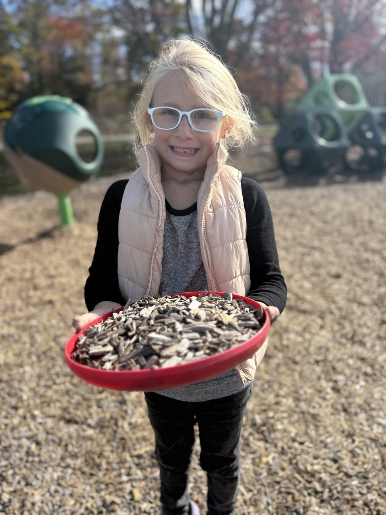 A child wearing glasses holds a red tray full of wood chips on the playground.