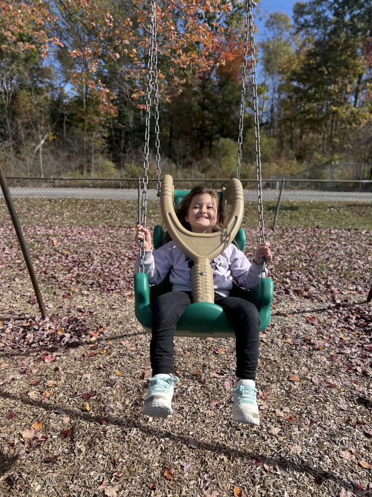 A smiling child swings in a green adaptive swing surrounded by autumn leaves.