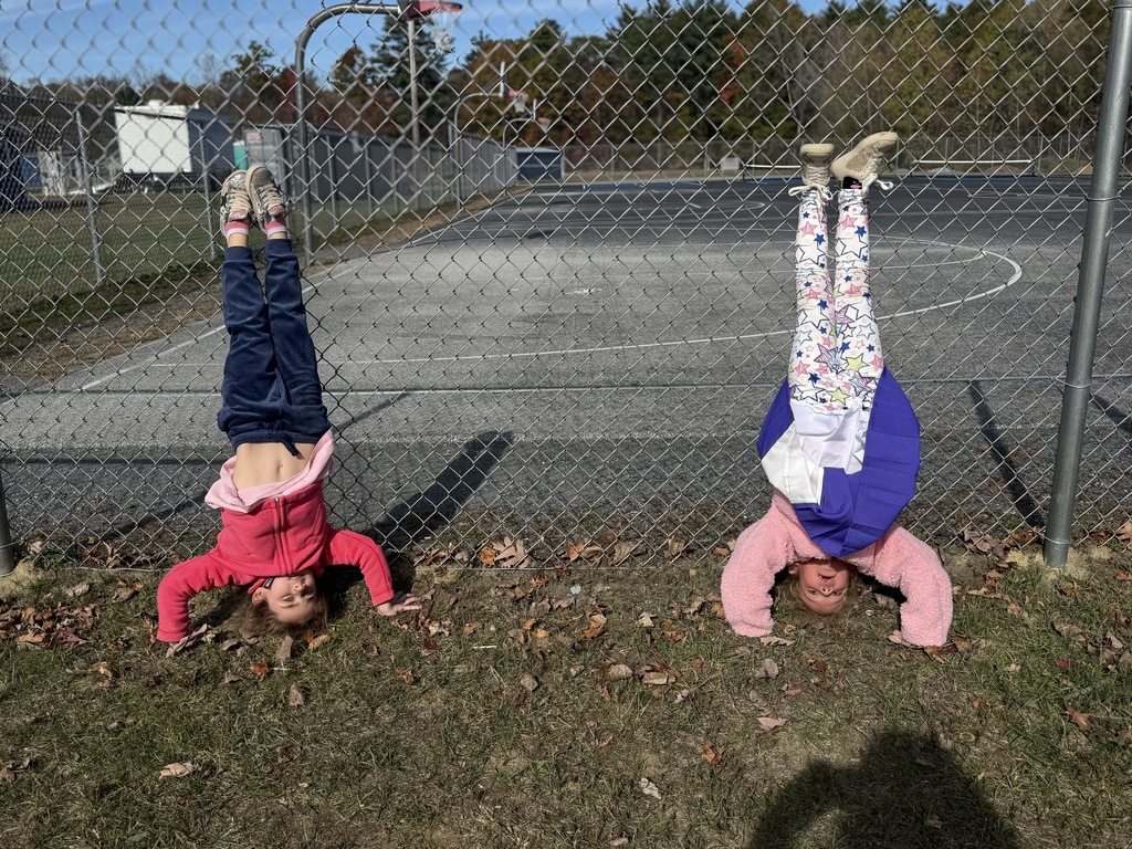Two children do handstands against a chain-link fence near a basketball court.