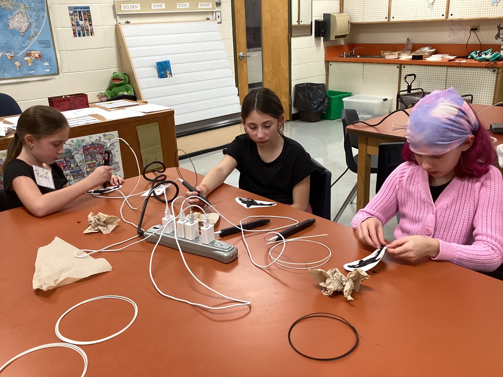 Three students using 3D pens at a classroom table filled with cords and supplies.