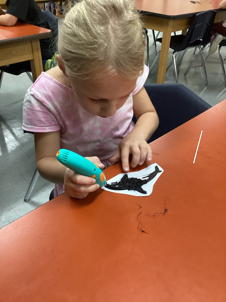Student using a 3D pen to fill in an orca whale outline at a classroom table.