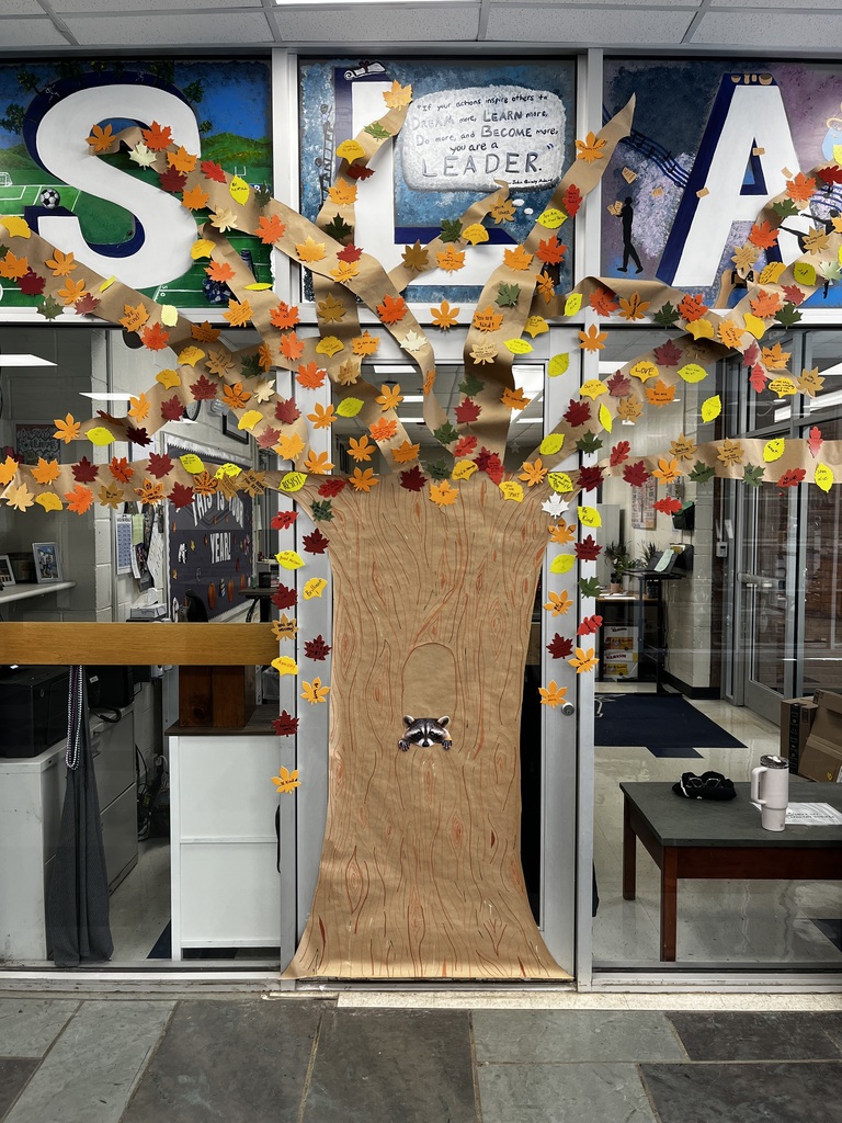 Completed “Kindness Tree” display made of brown paper with branches and leaves containing uplifting messages, set up in a school hallway with artwork spelling “SLATER” above it.