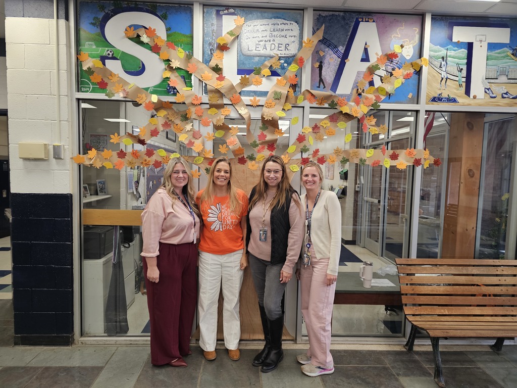 Four staff members stand smiling in front of a large paper tree decorated with colorful leaves featuring positive messages at the entrance of the school.