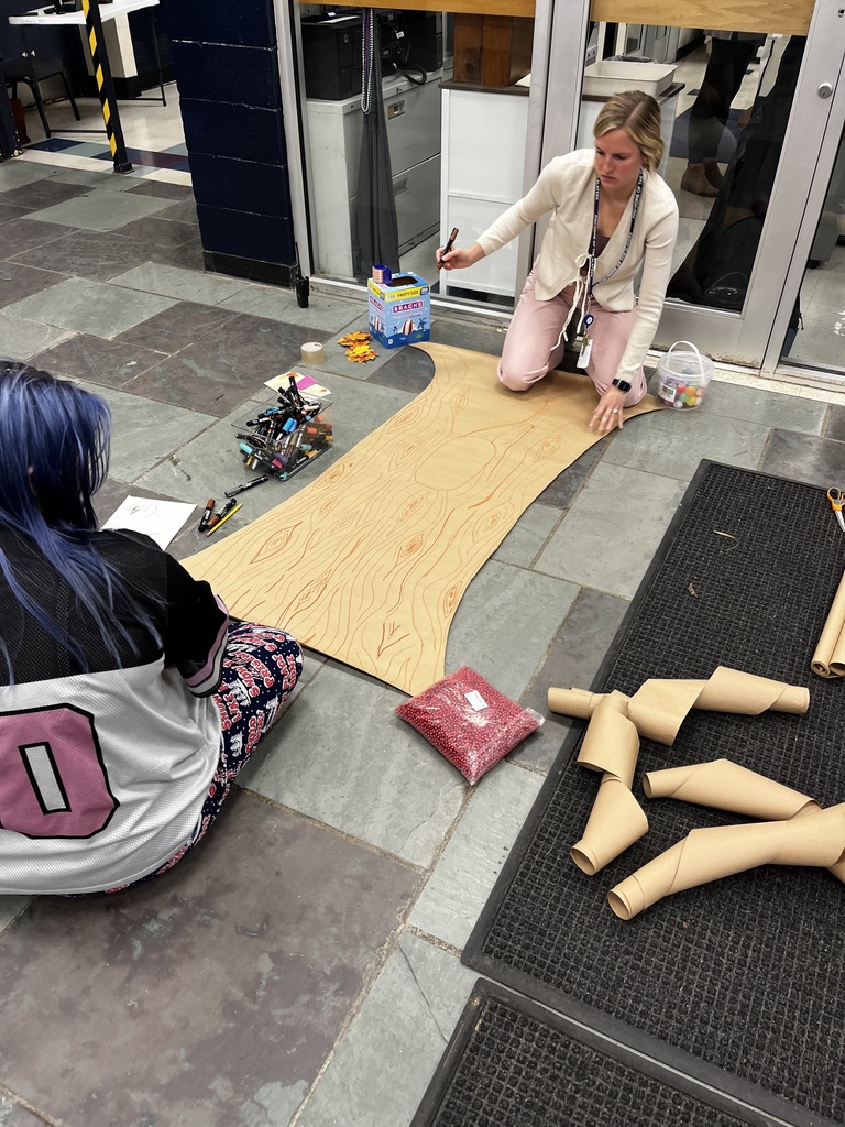 Two people work on drawing and assembling the base of a large paper tree on the floor, surrounded by markers, paper leaves, and supplies.