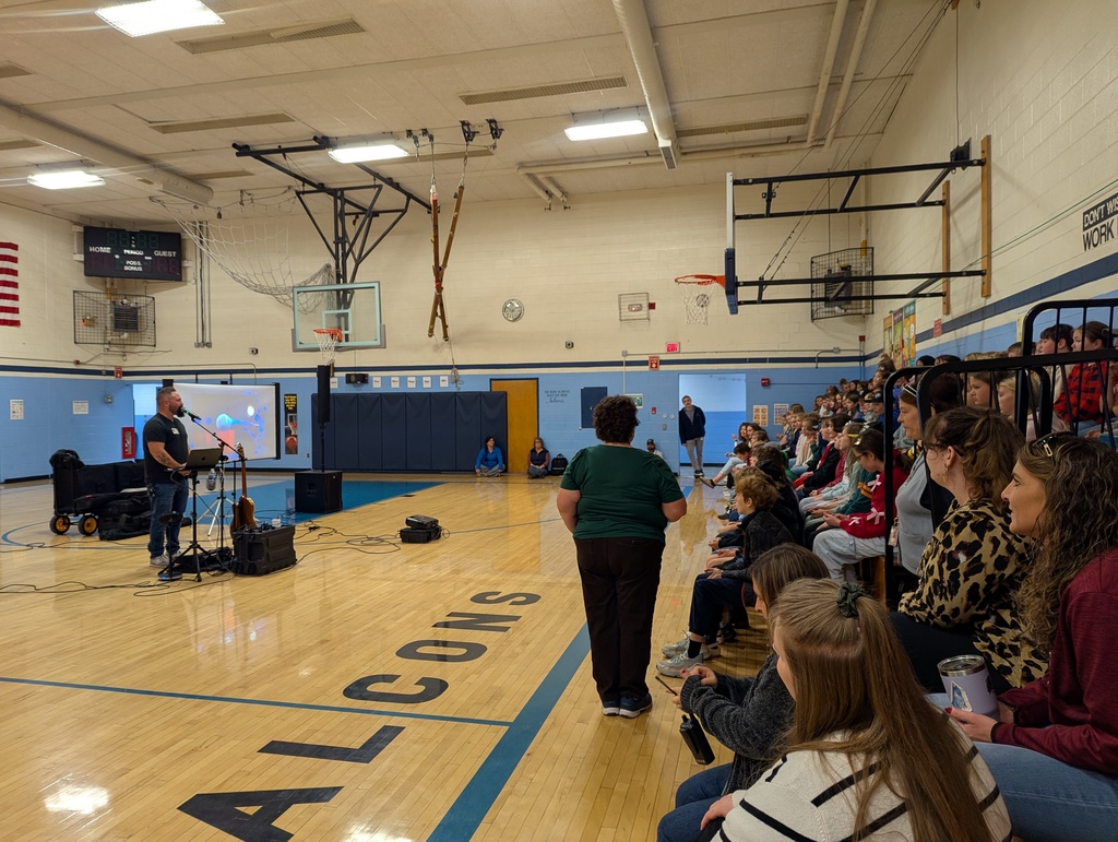 Musician performs in front of students and staff seated in bleachers, with a projector screen displaying visuals beside him.