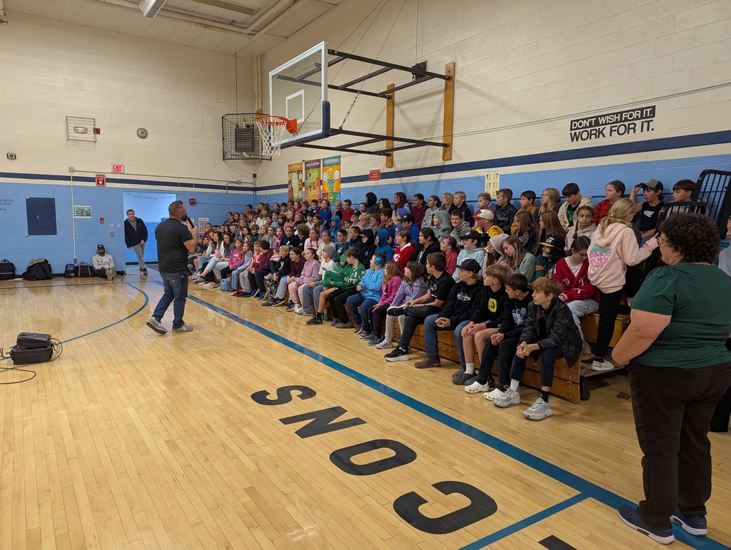 Musician speaks to a large group of older students sitting on bleachers in a school gym decorated with motivational posters.