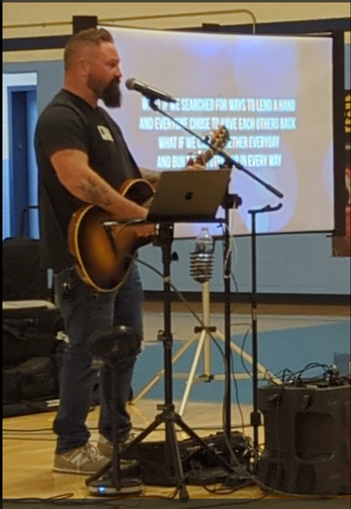 Close-up of musician playing guitar and singing into a microphone in the school gym, with lyrics projected on a screen behind him.