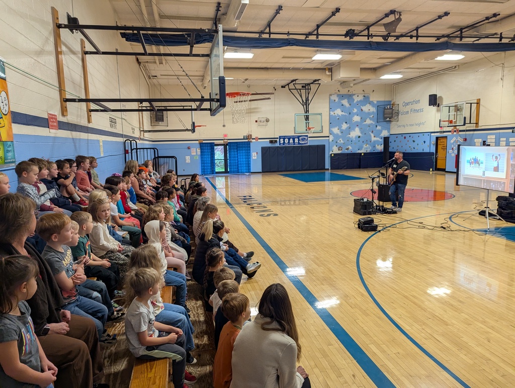 Elementary students seated on gym bleachers watch a musician perform with a guitar during a school assembly.