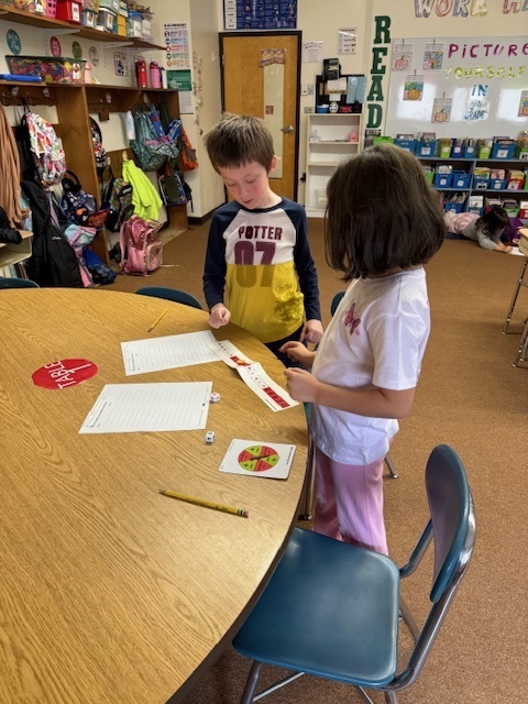 Two young students stand at a round wooden table in a classroom, engaged in a math activity. A boy in a Harry Potter shirt and a girl are looking down at papers, a ruler, a pair of dice, and a spinner on the table.
