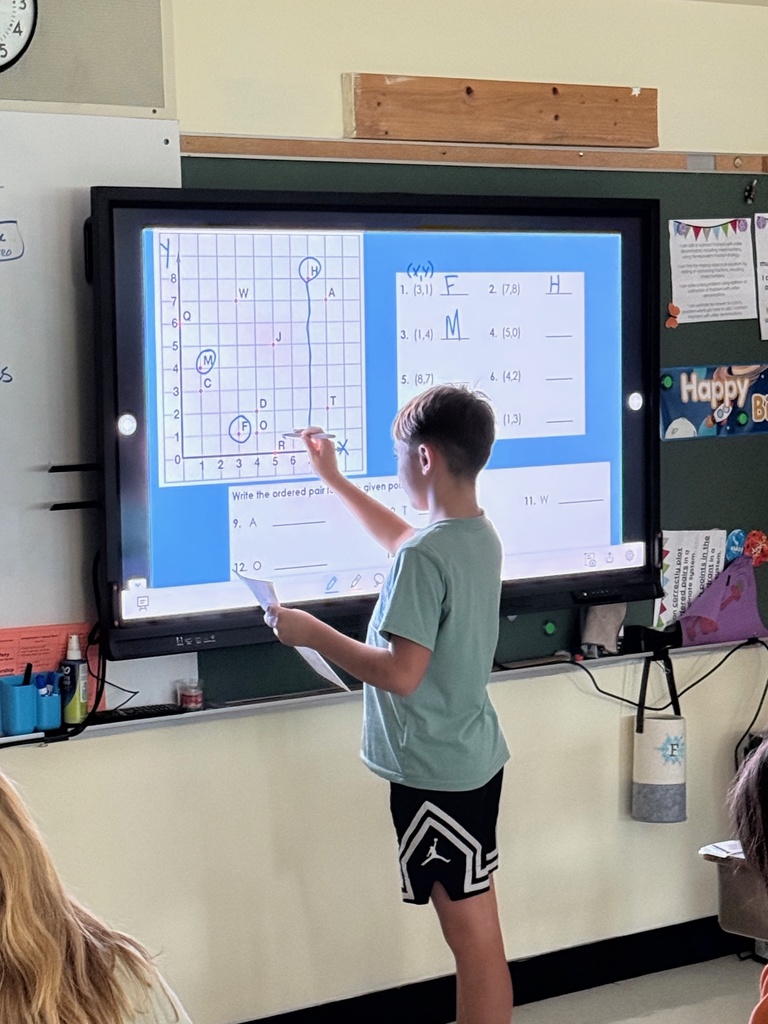 A student stands at a large interactive display in a classroom, pointing to a coordinate plane activity. The screen shows a grid with points plotted and labeled with letters, as well as questions about ordered pairs and points on the plane.