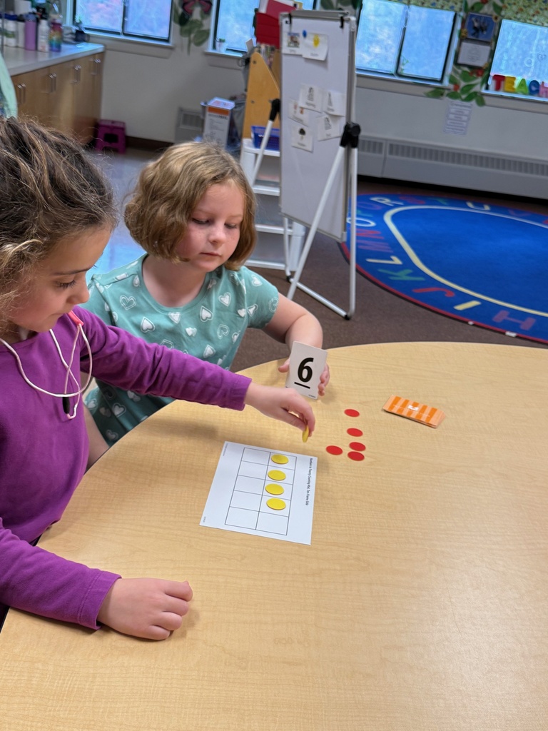 Two young girls sit at a table learning math. One holds a card with the number 6, and the other points to a ten-frame card where she is using yellow counters. There are also red counters on the table.