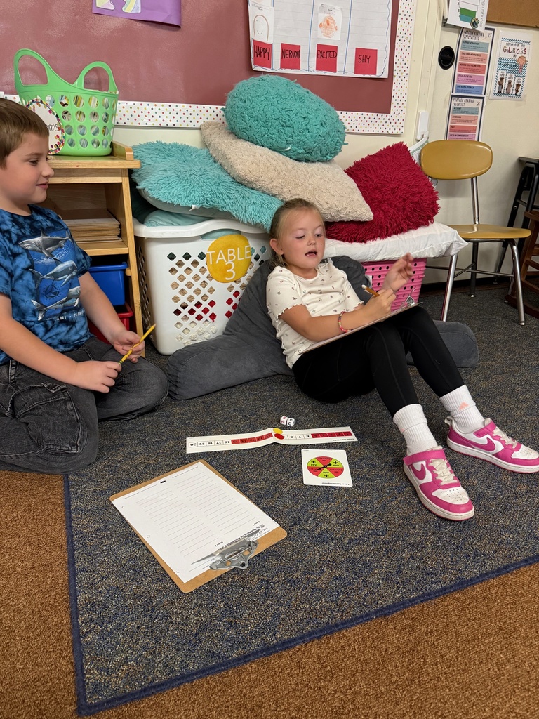 A young boy and girl sit on the floor in a classroom. The girl is leaning on a pillow in a reading nook, writing on a clipboard, while the boy sits nearby holding a pencil. A number line, dice, and a spinner are on the rug between them.