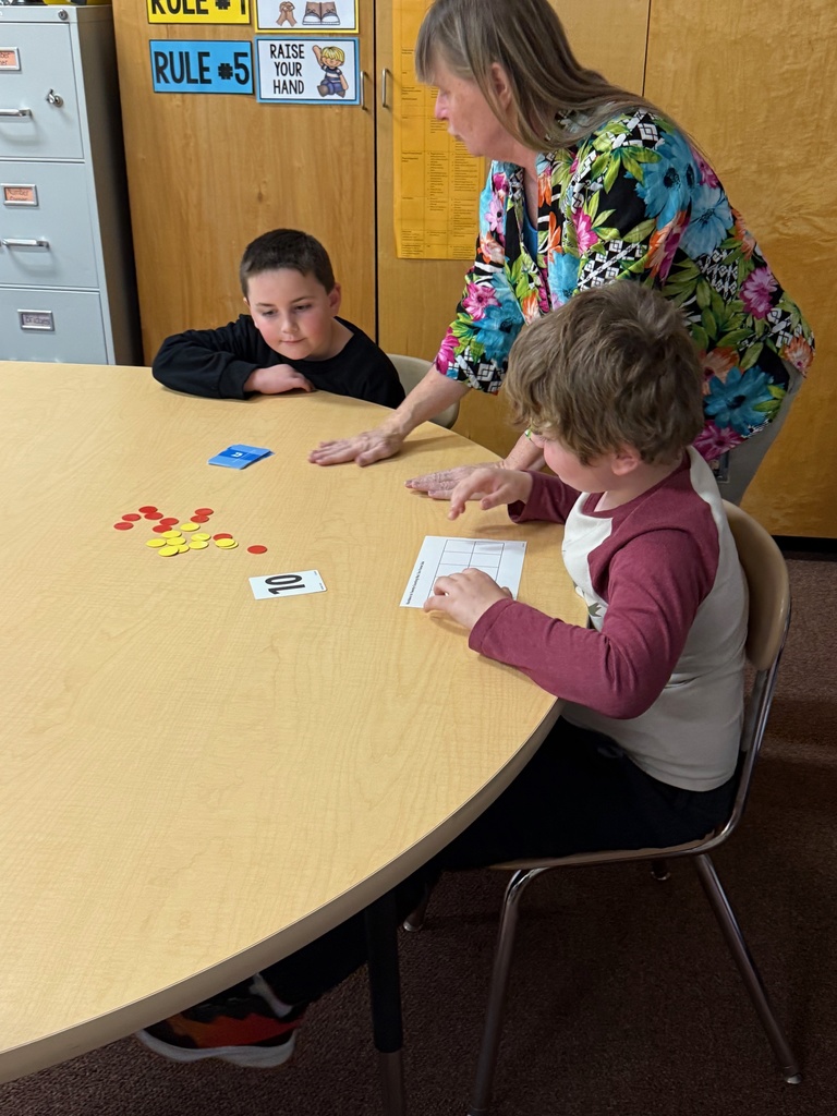 A teacher stands over two young male students sitting at a round wooden table, participating in a math lesson. The students are using a ten-frame card, number cards, and yellow and red counters. The teacher's hand is on the table near the materials.