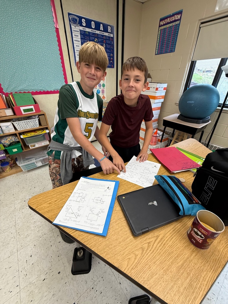 Two male students stand smiling behind a desk in a classroom, pointing at a paper with geometric figures on it. A Chromebook, a cup, a backpack, and other papers are on the desk with them.