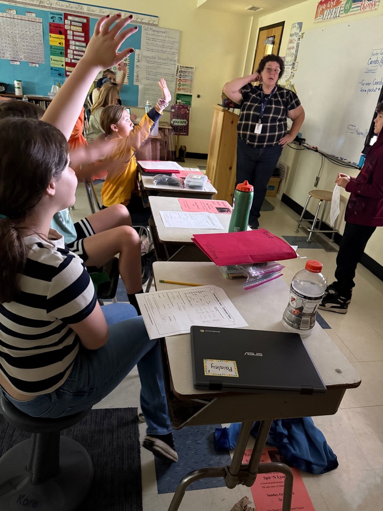 A middle-school classroom scene where students are actively participating; several students, including a girl in the foreground, have their hands raised. The teacher stands near a whiteboard in the background, observing the class. A Chromebook and papers are on the foreground student's desk.