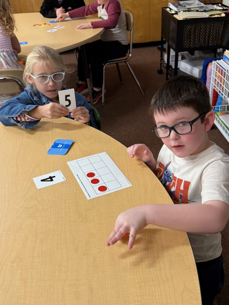 Two young students with glasses are sitting at a table engaged in a math activity. The girl is holding up a number card with a 5, and the boy is reaching for a red counter. On the table is a ten-frame card with four red counters, and a number card with a 4.