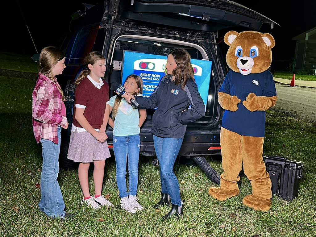 Three students are interviewed by an NBC 5 meteorologist outdoors at night beside the station’s weather vehicle, with a person in a bear mascot costume standing nearby.